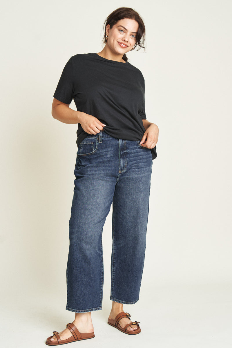 Side profile of plus-size model wearing dark blue curved leg jeans, showcasing the relaxed fit and comfortable design.